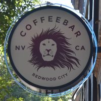 The circular sign from outside Coffeebar, a stylised lion's head with flowing mane, the words "Coffeebar" at the top and "Redwood City" at the bottom, flanked by the letters NV (Nevada, left) and CA (California, right)