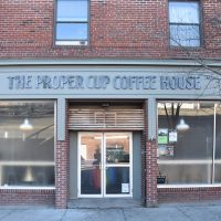 The front of The Proper Cup on Forest Avenue, Portland, the central double doors flanked by a pair of windows with the words "The Proper Cup Coffee House" written across the top.